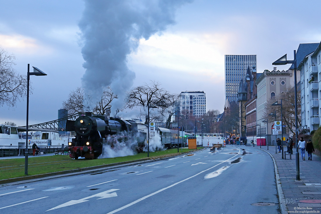 Auf der Hafenbahn in Frankfurt am Main werden von der „Historischen Eisenbahn Frankfurt“ regelmäßig Sonderfahrten veranstaltet. Mit der vereinseigenen Dampflok 52 4867, die hier in der Mainmetropole irgendwie wie ein Fremdkörper wirkt, verlässt der Zug den Haltepunkt Eiserner Steg, 14.12.2019.